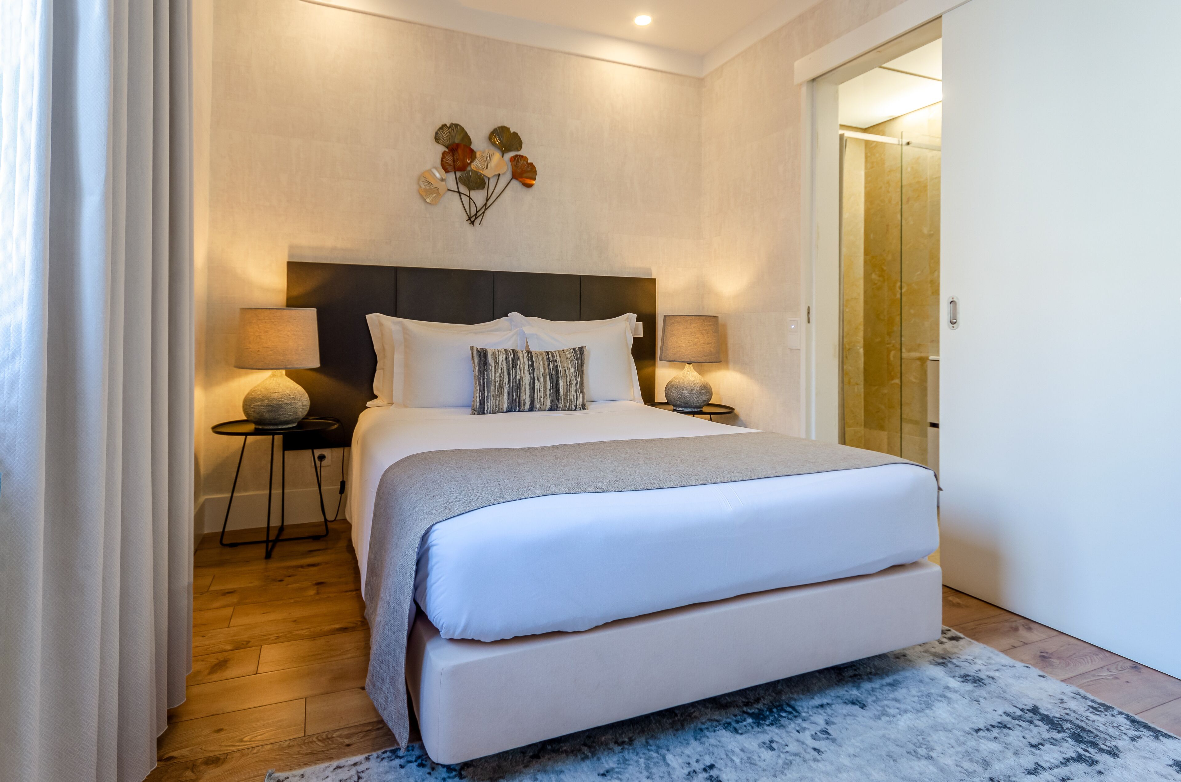 A modern bedroom with a large bed, two bedside tables with lamps, a decorative wall piece, and a wooden floor.