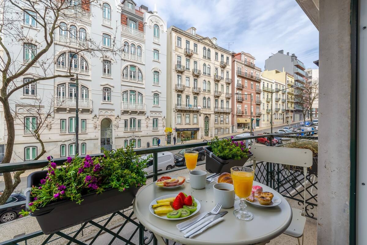 A balcony with a table set for breakfast, featuring a plate of fruit, a croissant, and a glass of orange juice.