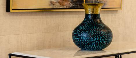 A blue vase with a textured pattern on a table against a wooden wall with a framed painting.