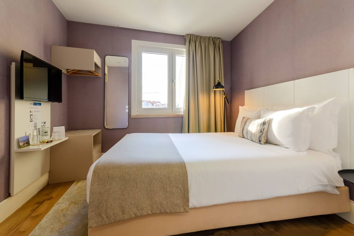A hotel room with a large bed, a TV mounted on the wall, a window with curtains, and a bedside table with a lamp.