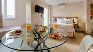Habitación de hotel con mesa de comedor de vidrio, bufé de desayuno, cama y televisor montado en la pared.