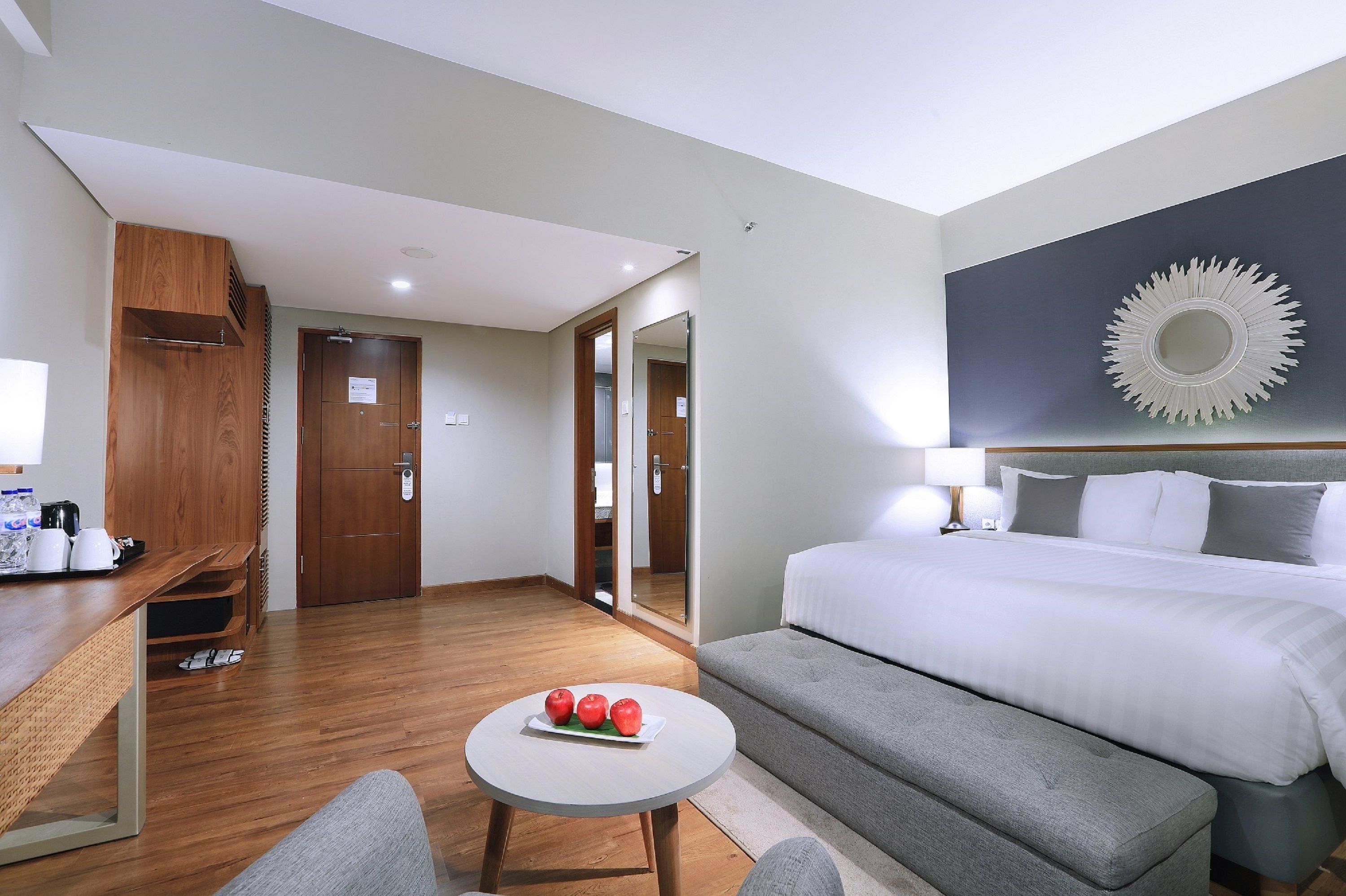 Habitación de hotel con cama, escritorio con laptop, pequeña mesa redonda con manzanas y sofá gris.