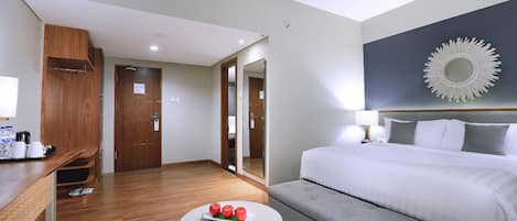 Habitación de hotel con cama, escritorio con laptop, pequeña mesa redonda con manzanas y sofá gris.