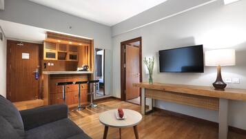 A modern living room with a sofa, a small round table with a decorative item, a kitchen area with a bar stool, a television mounted on the wall, and a wooden bench with a lamp.