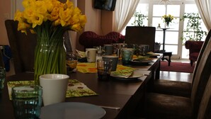 Een eetruimte met een tafel gedekt voor twee personen, een vaas met gele bloemen en een aan de muur bevestigde televisie.
