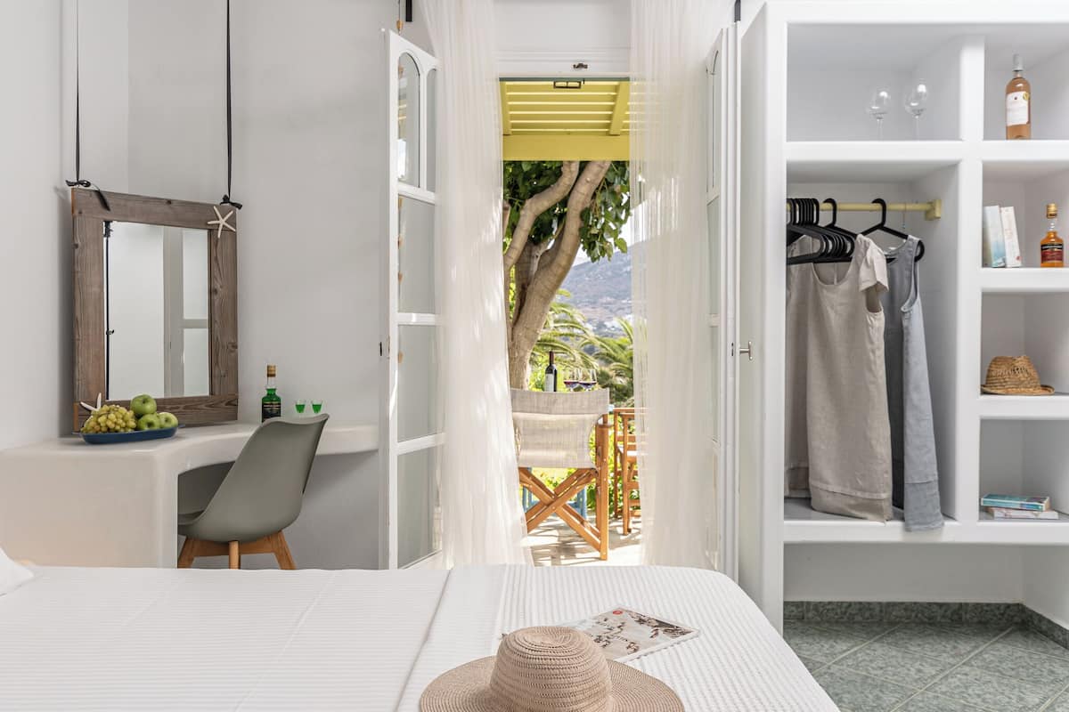 Ein Schlafzimmer mit Bett, Stuhl, Schreibtisch mit Spiegel, Kleiderschrank und Blick ins Freie.