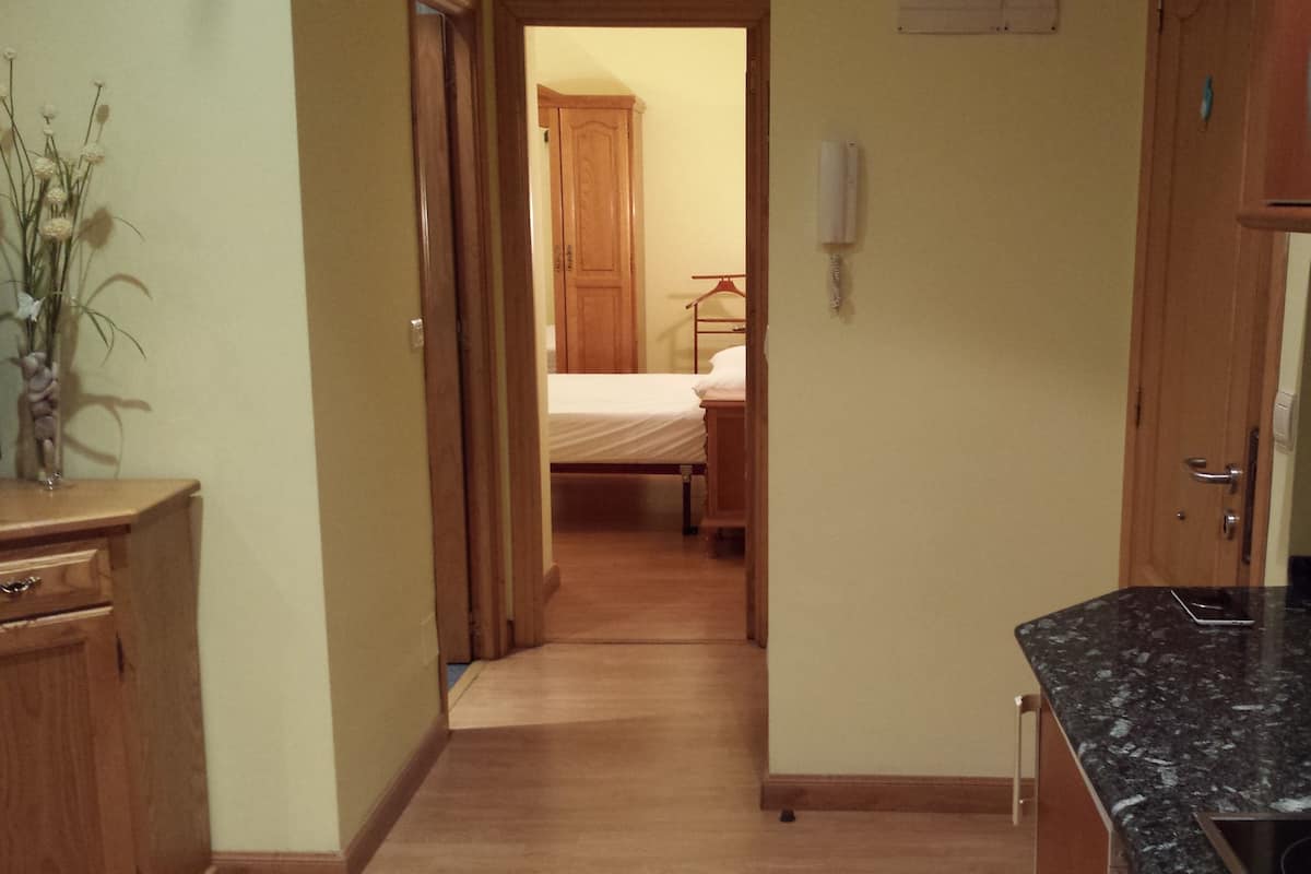 A narrow hallway leading to a bedroom with a wooden bed and a bedside table. There is a wooden dresser on the left and a kitchen area with a granite countertop on the right.