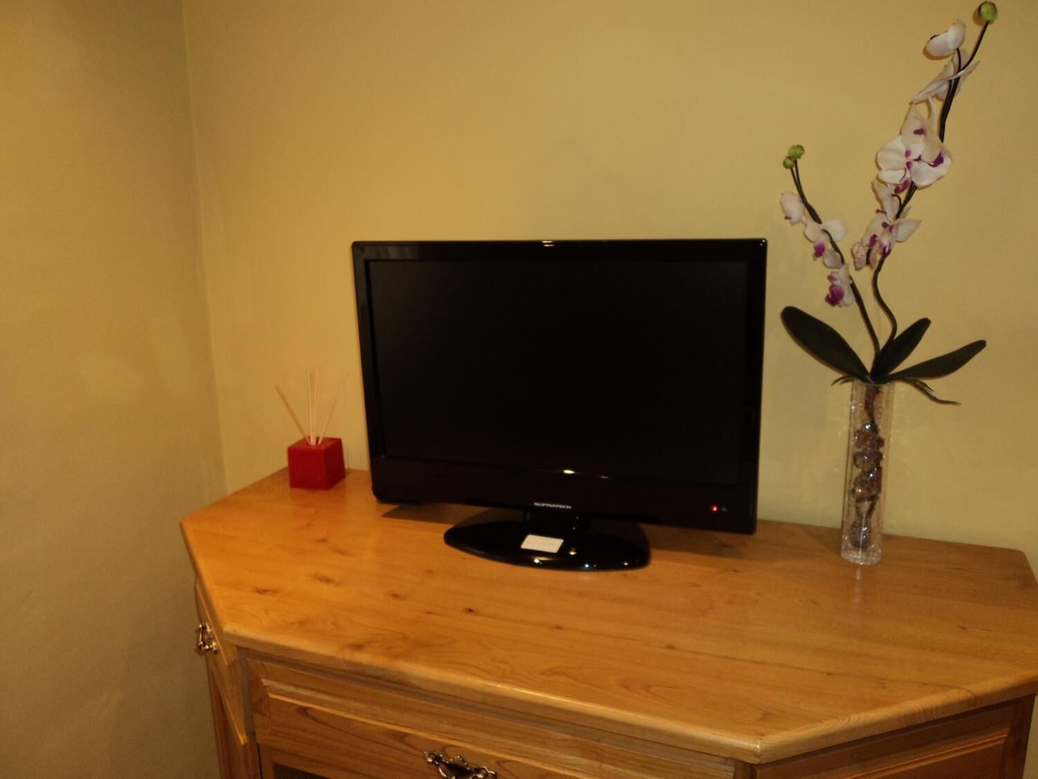 Un meuble en bois pour la télévision, sur lequel est posée une télévision à écran plat, un porte-bougie rouge et un vase contenant des orchidées.