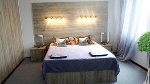 Quarto moderno com cabeceira de madeira, uma cama grande com colcha azul, duas mesas de cabeceira com luminárias e um vaso com planta.