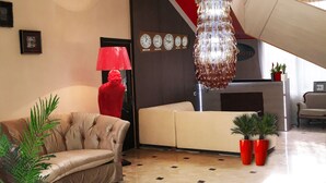 Um hall de entrada com um candelabro, uma luminária vermelha, um sofá, duas plantas em vasos e um balcão de recepção.