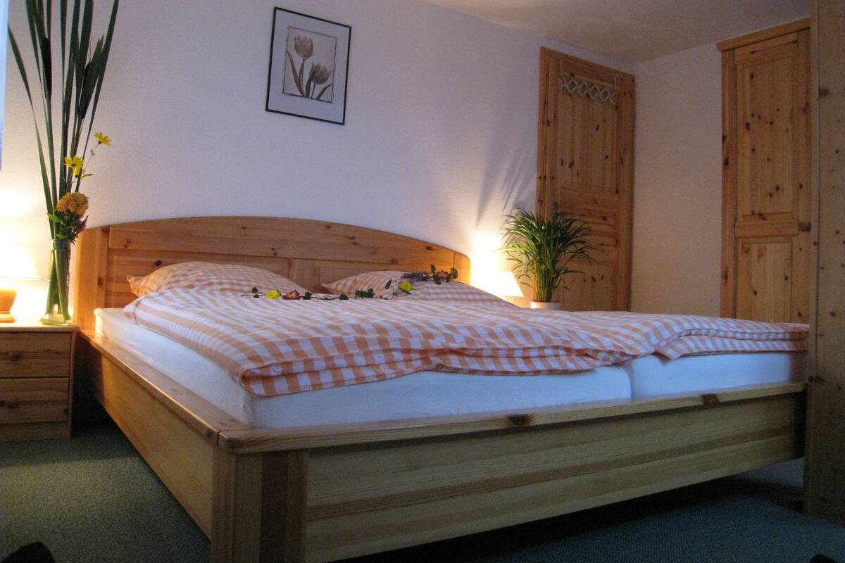 A bedroom with a wooden bed, a nightstand with a lamp, a potted plant, and a wooden wardrobe.