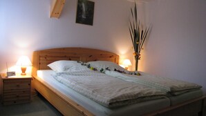 A bedroom with a wooden bed, two bedside tables with lamps, a nightstand, a wall-mounted picture, and a tall vase with flowers. - Kammbaude Neuhermsdorf (Hermsdorf-Erzgebirge)