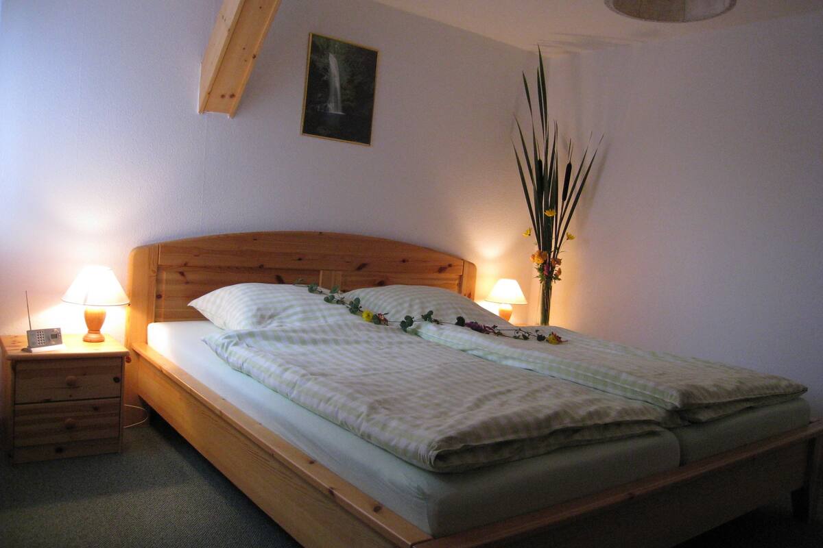 A bedroom with a wooden bed, two bedside tables with lamps, a nightstand, a wall-mounted picture, and a tall vase with flowers.