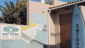 Entrada a una escuela de surf con un mural, una tabla de surf y un gato sentado cerca.