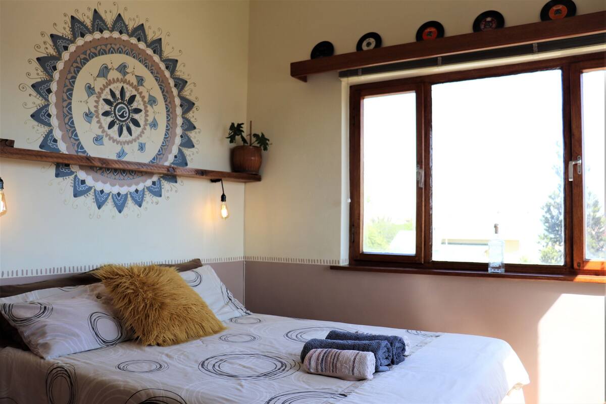 Ein Schlafzimmer mit einem Bett, einer großen Wandteppich, einem Fenster mit Jalousien und einem Regal mit dekorativen Gegenständen.