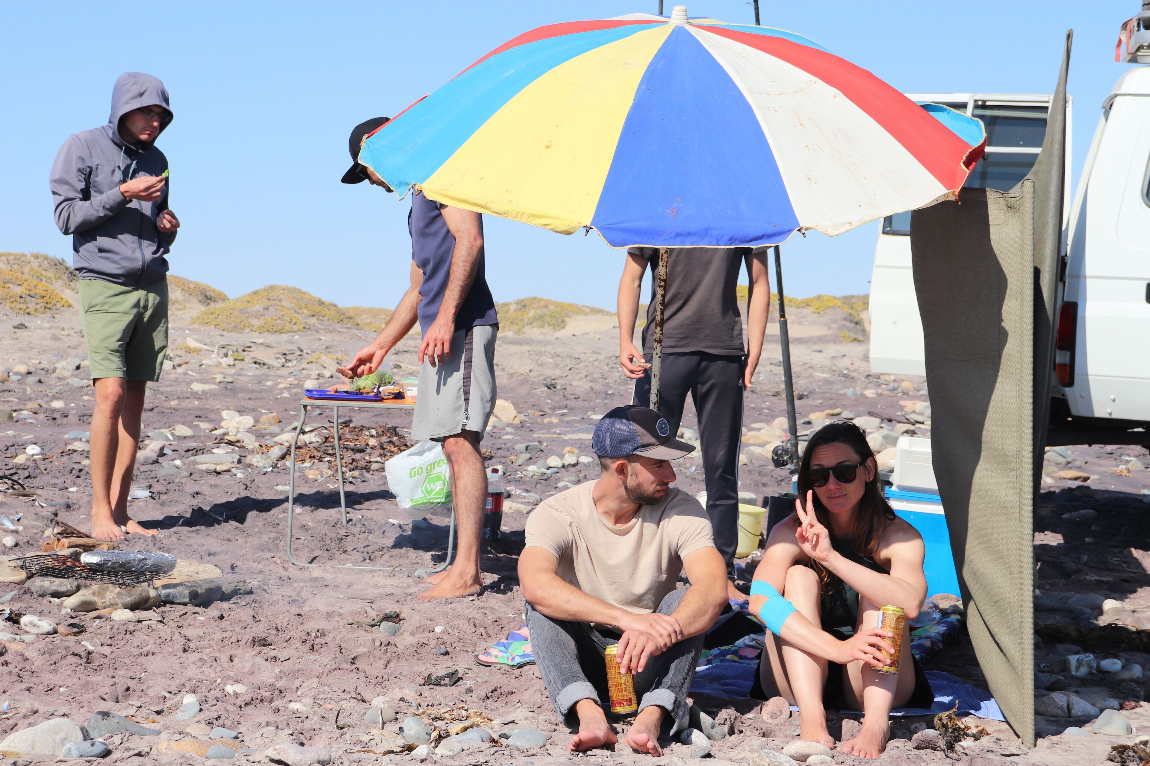 A group of people sitting under a colorful umbrella on a rocky beach with a white vehicle in the background.
