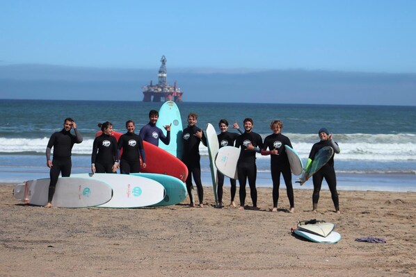 Un groupe de personnes, équipées de planches de surf, se trouve sur une plage, avec une plateforme pétrolière en mer en arrière-plan.