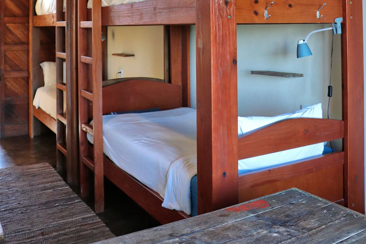 Ein schlichtes Zimmer mit einem Holzbettgestell, einer Klapptreppe, einem kleinen Tisch und einer Hängelampe.