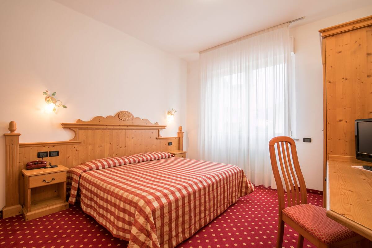 Quarto de hotel com cama de madeira, mesa de cabeceira de madeira, cadeira de madeira, televisão e janela com cortinas brancas.