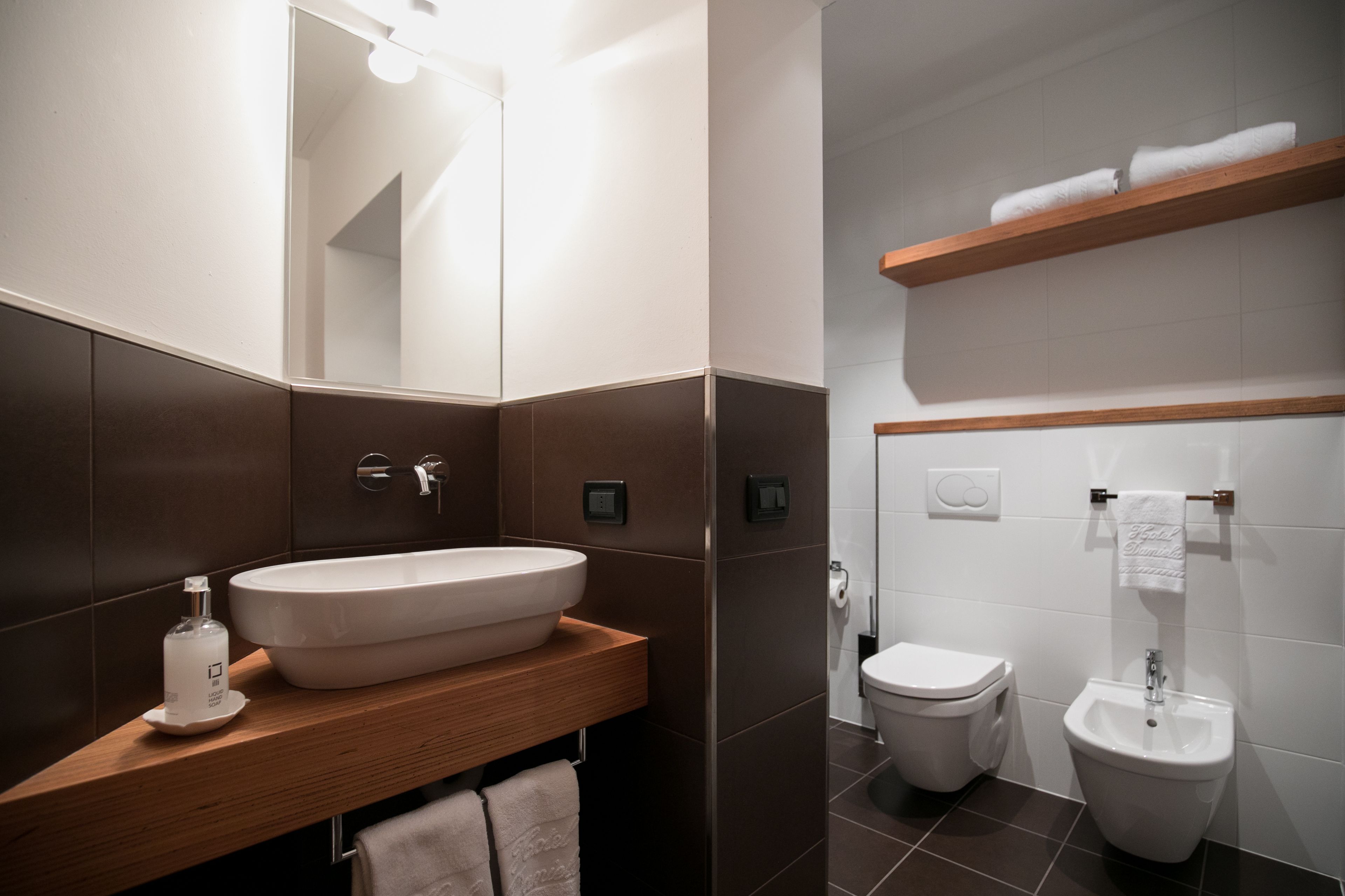 Un bagno moderno con un ampio lavandino ovale, un water e un bidet. Sopra il water, uno scaffale in legno porta degli asciugamani.