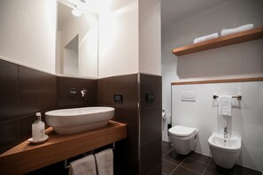 A modern bathroom with a large oval sink, a toilet, and a bidet. There is a wooden shelf above the toilet holding towels. - Hotel Daniela (Levico Terme)
