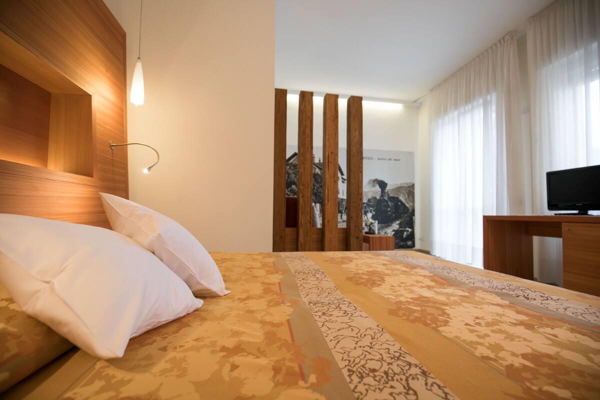 Quarto de hotel moderno com cabeceira de madeira, cama grande com roupa de cama estampada, escrivaninha com televisão de tela plana e vista para uma paisagem montanhosa através de cortinas leves.