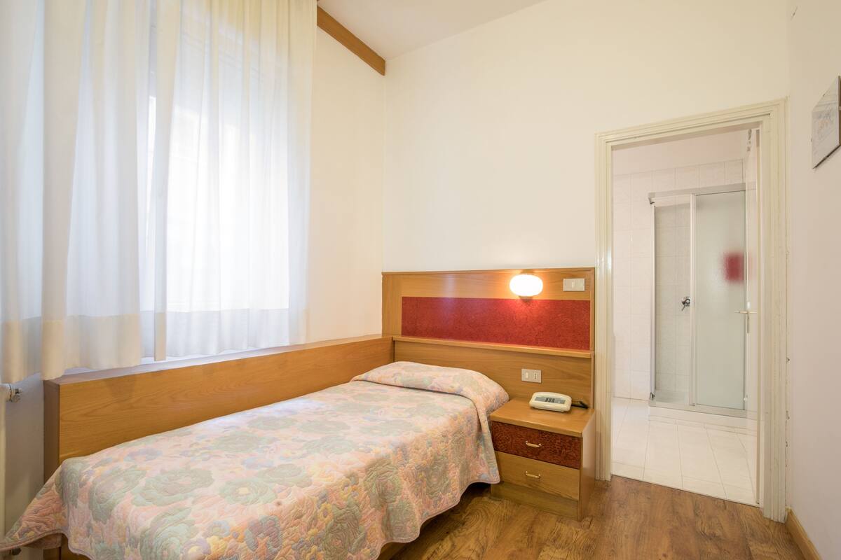 Um quarto pequeno e bem arrumado com uma cama de solteiro, uma mesa de cabeceira com abajur, e uma porta que dá para o banheiro.