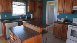 A kitchen with wooden cabinets, a built-in island, a dishwasher, a stove, and a window with a patterned valance. - Seaside Nest on Private Waterfront (Chéticamp)