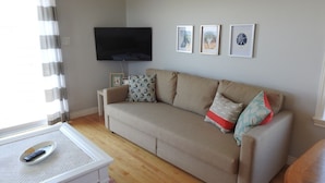 A living room with a beige sofa, a flat-screen TV mounted on the wall, and three framed pictures. - Seaside Nest on Private Waterfront (Chéticamp)