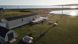 A mobile home with a deck and a small shed, situated near a body of water with a clear sky. - Seaside Nest on Private Waterfront (Chéticamp)