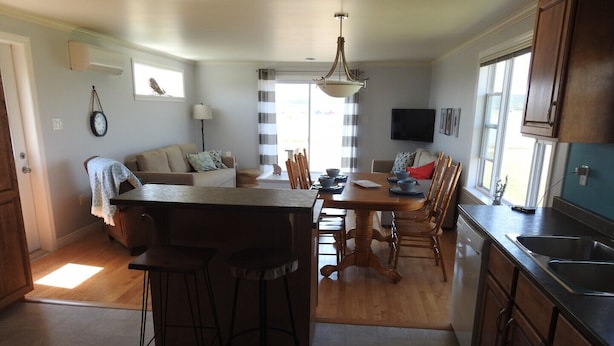 A modern kitchen with a central island, bar stools, a dining table with chairs, a TV mounted on the wall, and a large window providing natural light. - Seaside Nest on Private Waterfront (Chéticamp)