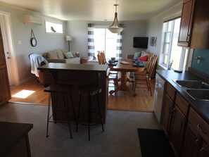 A modern kitchen with a central island, bar stools, a dining table with chairs, a TV mounted on the wall, and a large window providing natural light. - Seaside Nest on Private Waterfront (Chéticamp)