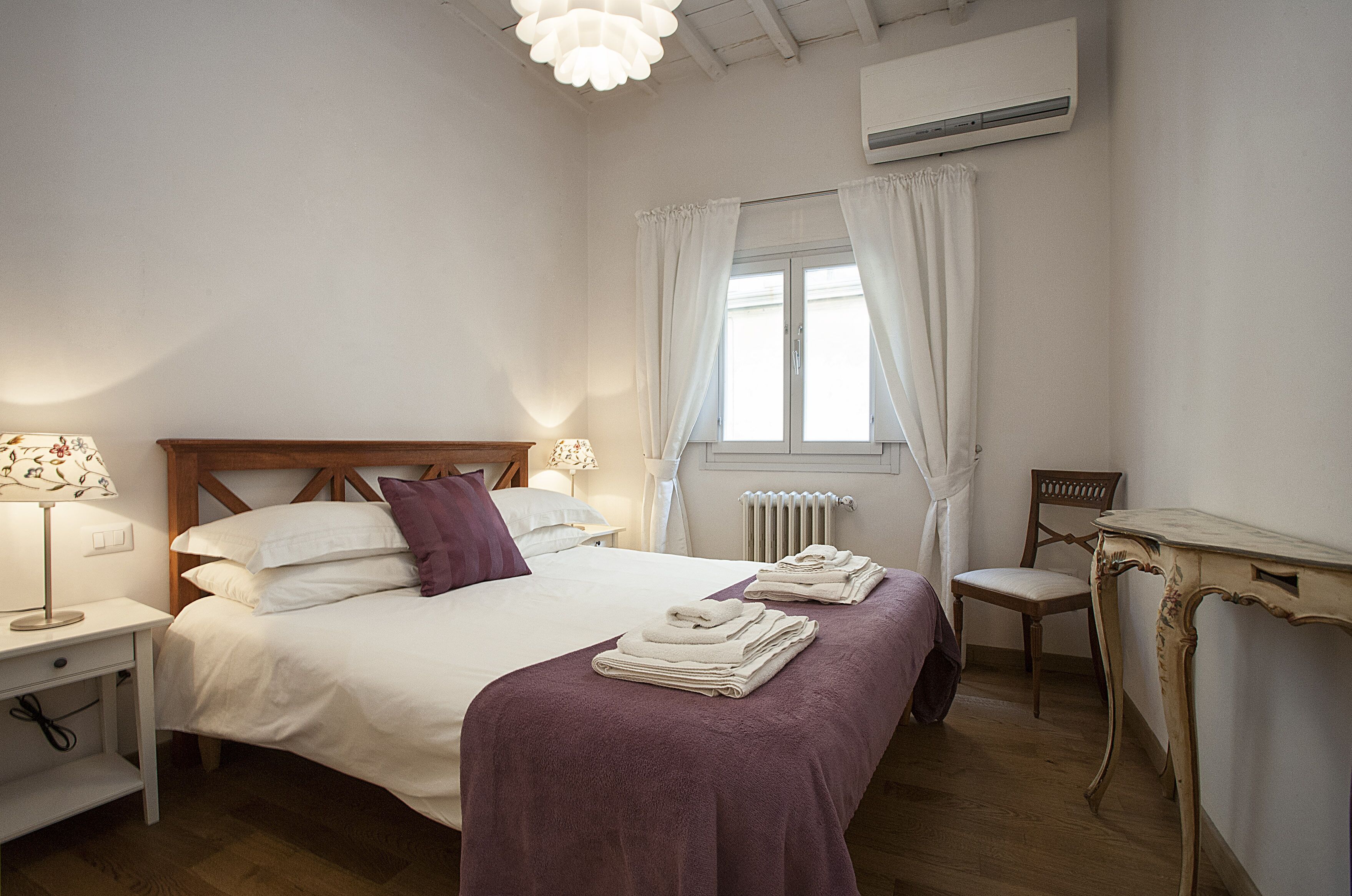 A neatly made bed with white and purple bedding, a wooden headboard, and a bedside table with a lamp.