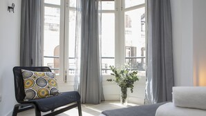 Ein Hotelzimmer mit Bett, einem Stuhl mit gemustertem Kissen, einer Vase mit Blumen und Blick auf Gebäude durch große Fenster.