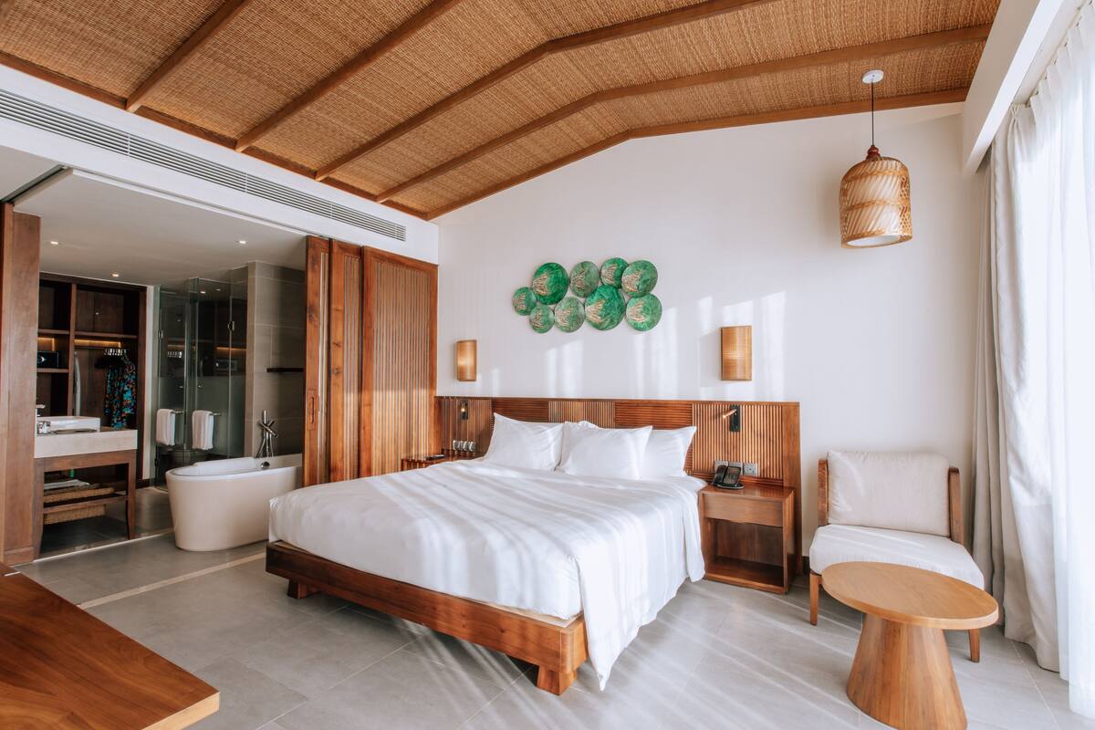 A modern bedroom with a large bed, wooden headboard, a round wooden table, a chair, a wall-mounted light fixture, and a bathroom with a glass shower enclosure.