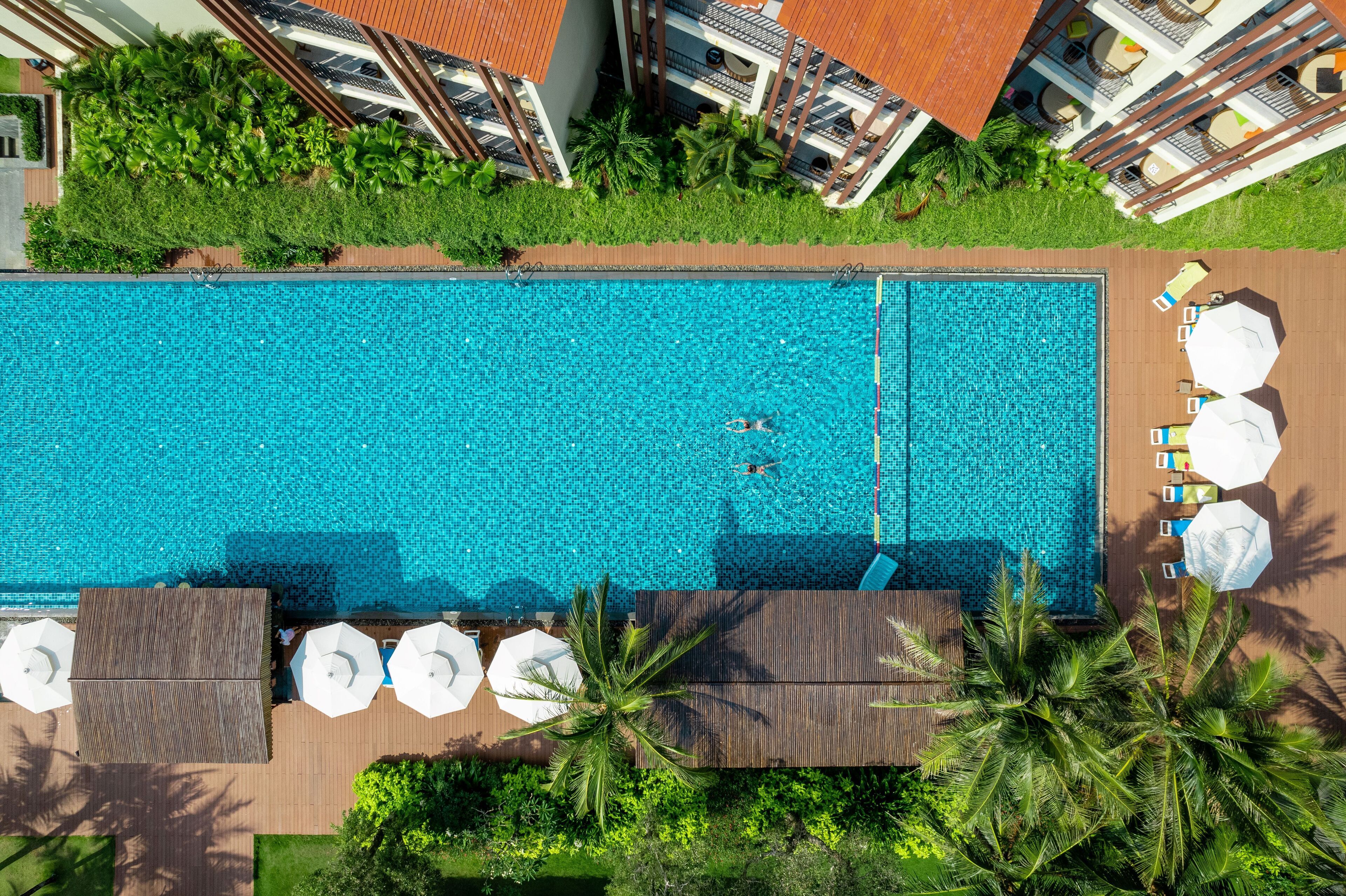 Vista aérea de una piscina con sillas de descanso y sombrillas, rodeada de follaje y edificios.