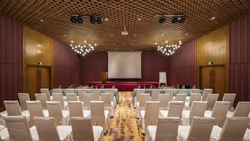 A conference room with rows of white chairs, a stage with a red carpet, and a large screen.