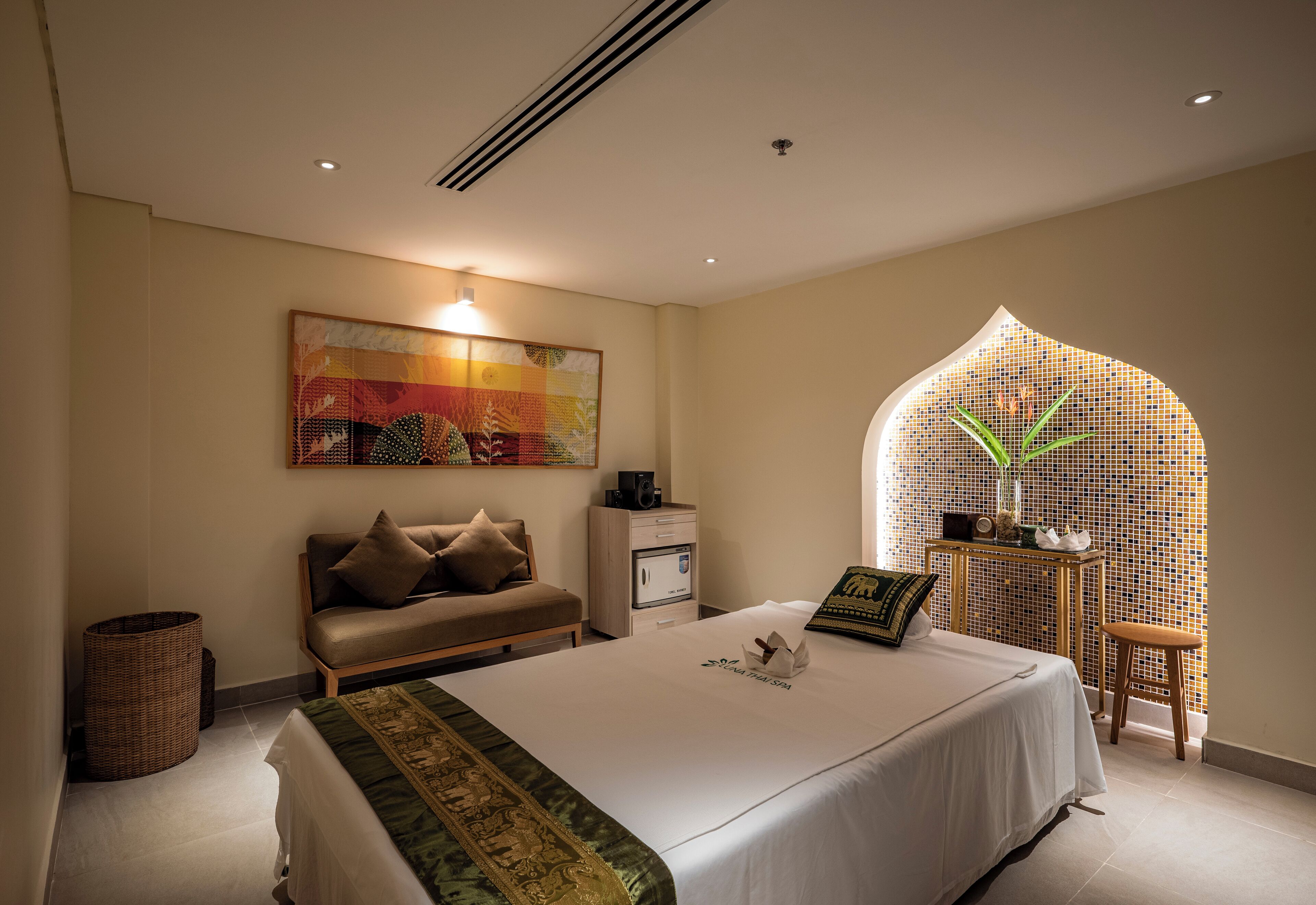 A hotel room with a bed, a sofa, a small table, a wall-mounted TV, and a window with a decorative arch.