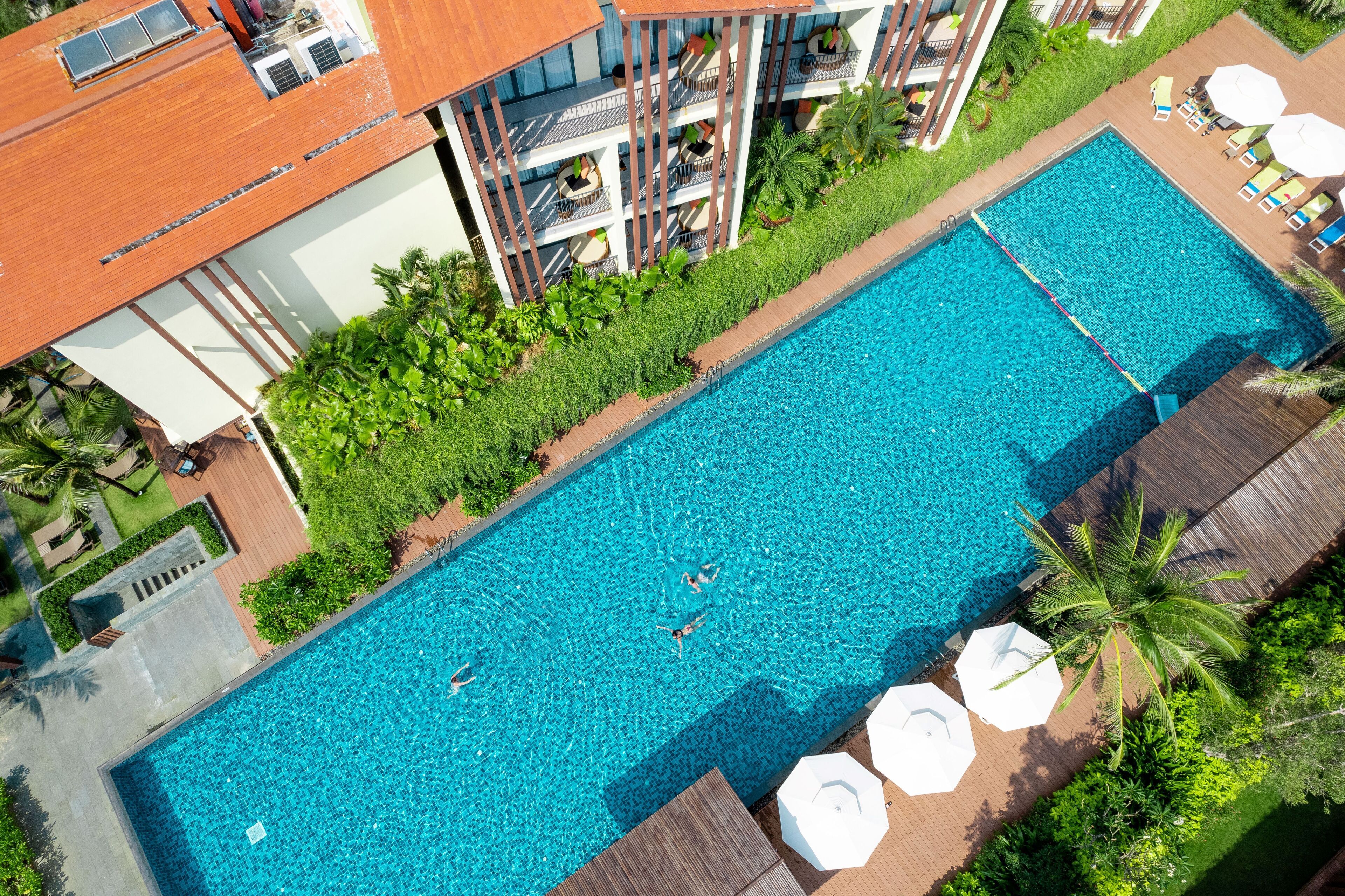 An aerial view of a hotel with a rectangular swimming pool surrounded by lush greenery, a multi-story building with balconies, and outdoor seating areas.