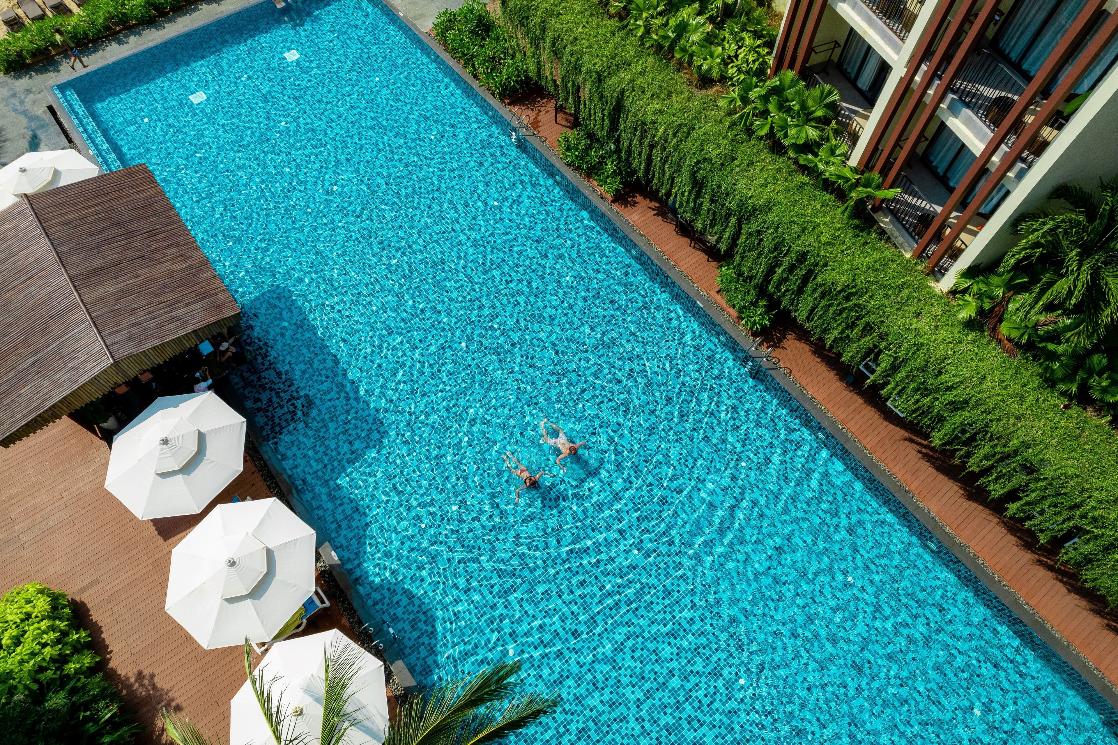 Vista aérea de una piscina rectangular con dos personas nadando, rodeada de abundante vegetación y una zona de terraza de madera con sombrillas.