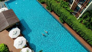 Vista aérea de una piscina rectangular con dos personas nadando, rodeada de abundante vegetación y una zona de terraza de madera con sombrillas.