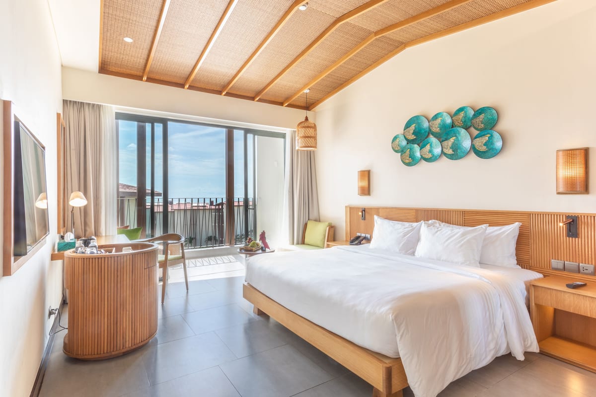 A modern hotel room with a large bed, a wooden headboard, a round wooden side table, a large window with curtains, a balcony, and a wall-mounted teal decorative piece.