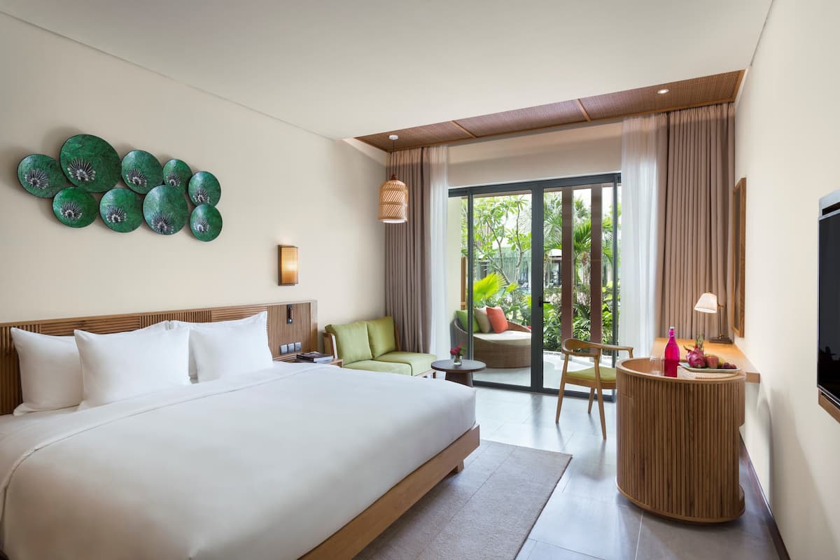 A modern hotel room with a large bed, a seating area with a green sofa and armchairs, a small table with a lamp, and a wall-mounted decorative piece.