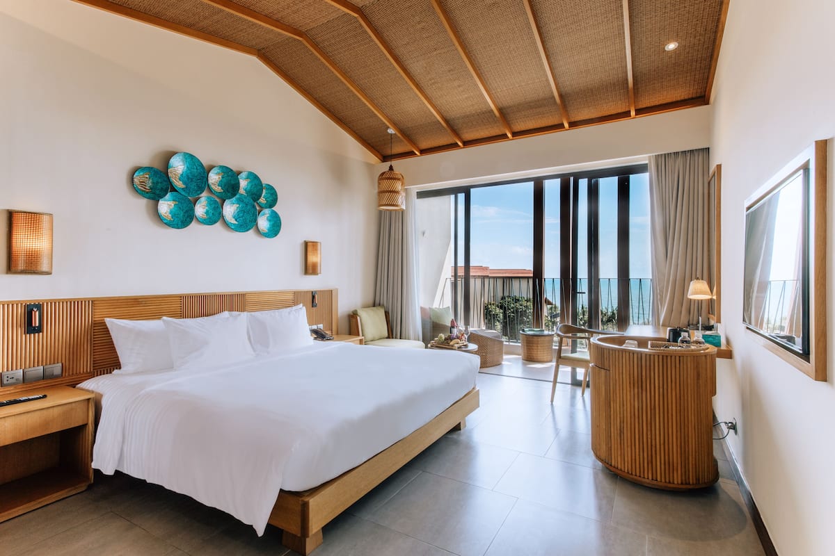 A modern hotel room with a large bed, a wooden headboard, a round wooden side table, and a balcony with a view.