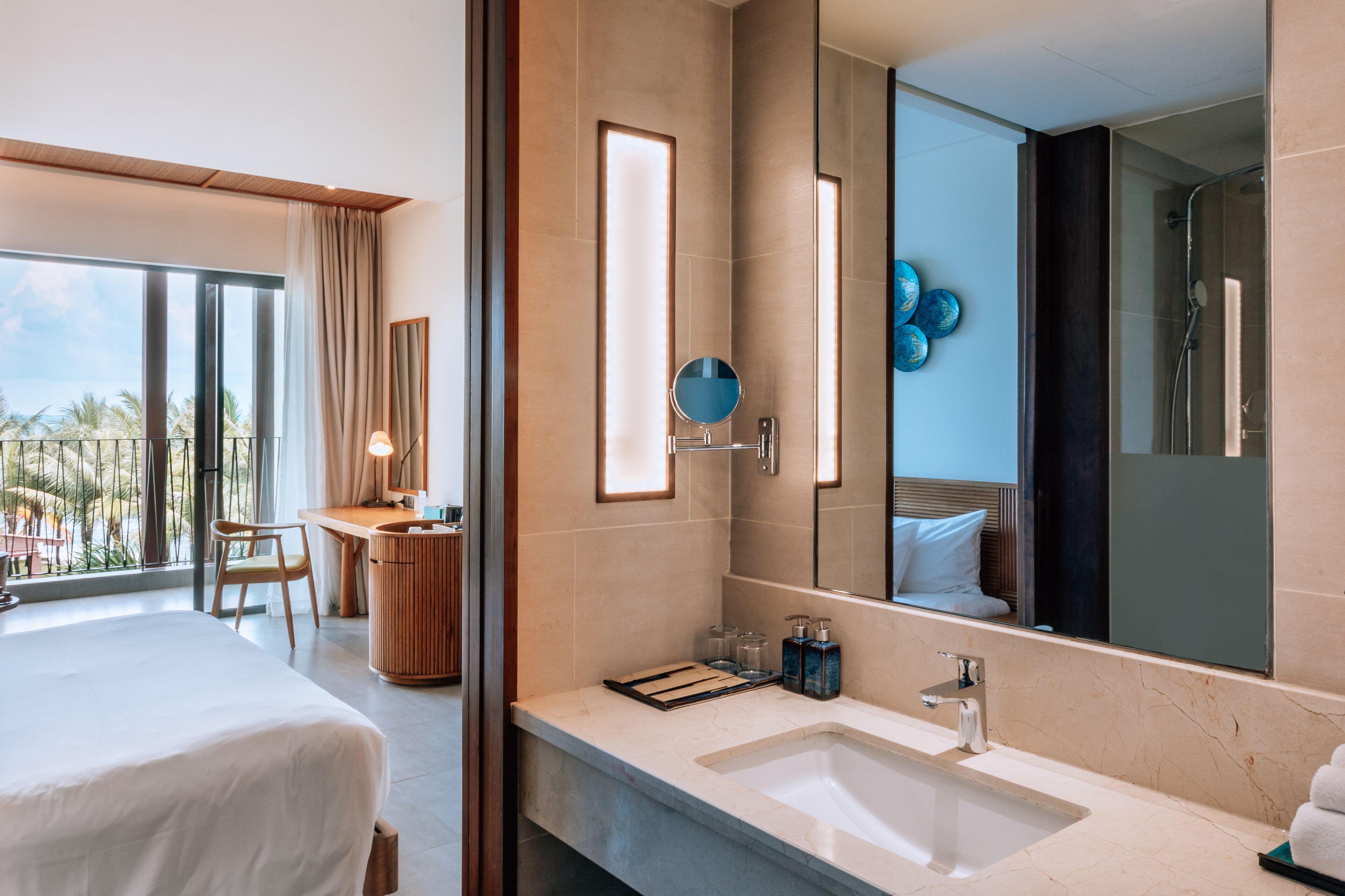 A modern hotel room with a large bed, a desk, a bathroom with a large mirror, and a view of palm trees.