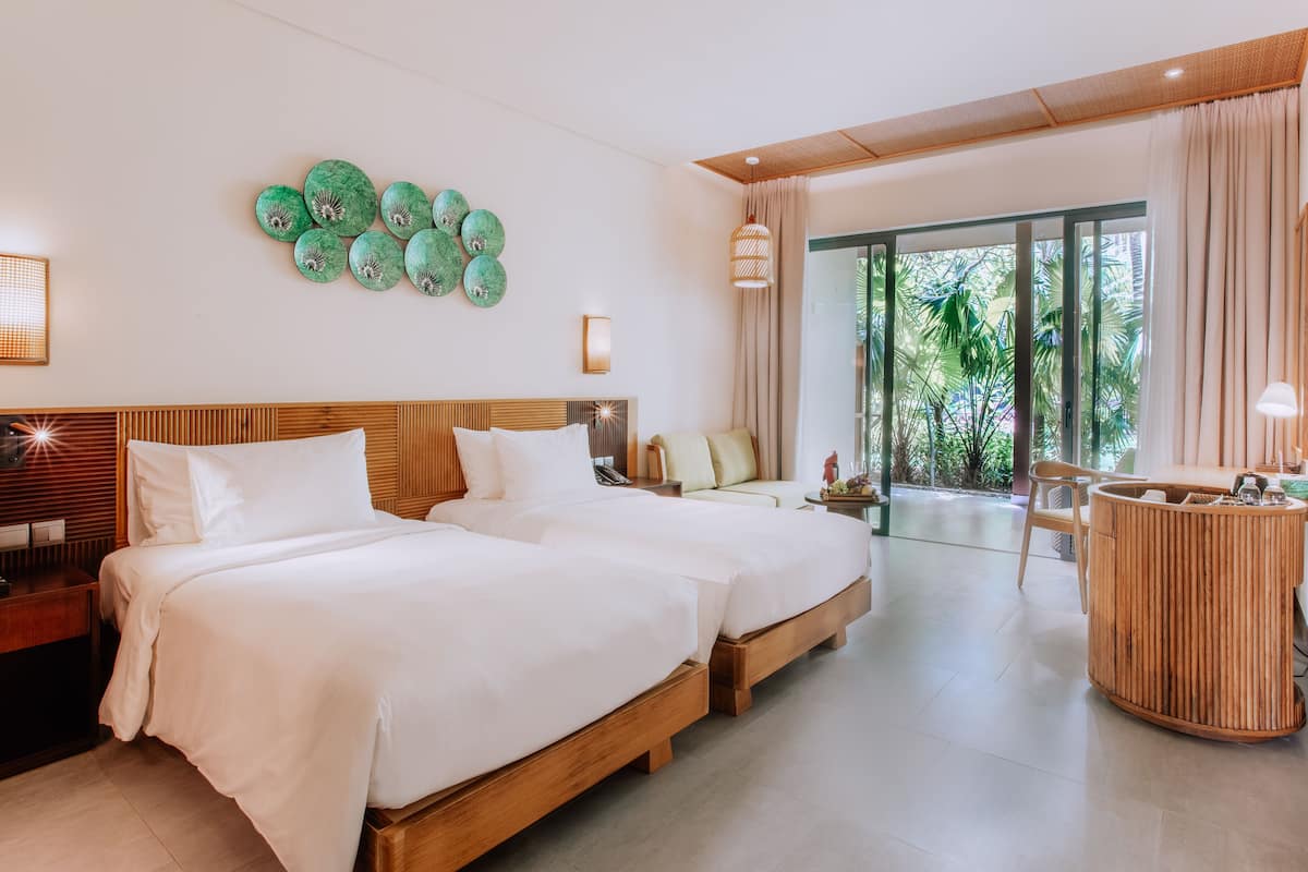 A modern hotel room with two beds, a wooden headboard, a round wooden side table, and a large window with a view of greenery.