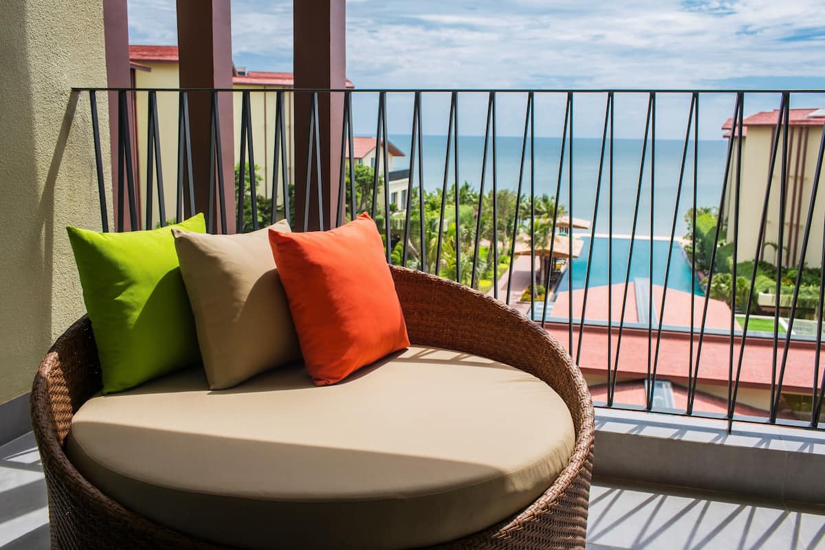 A wicker chair with cushions on a balcony overlooking a pool and ocean.
