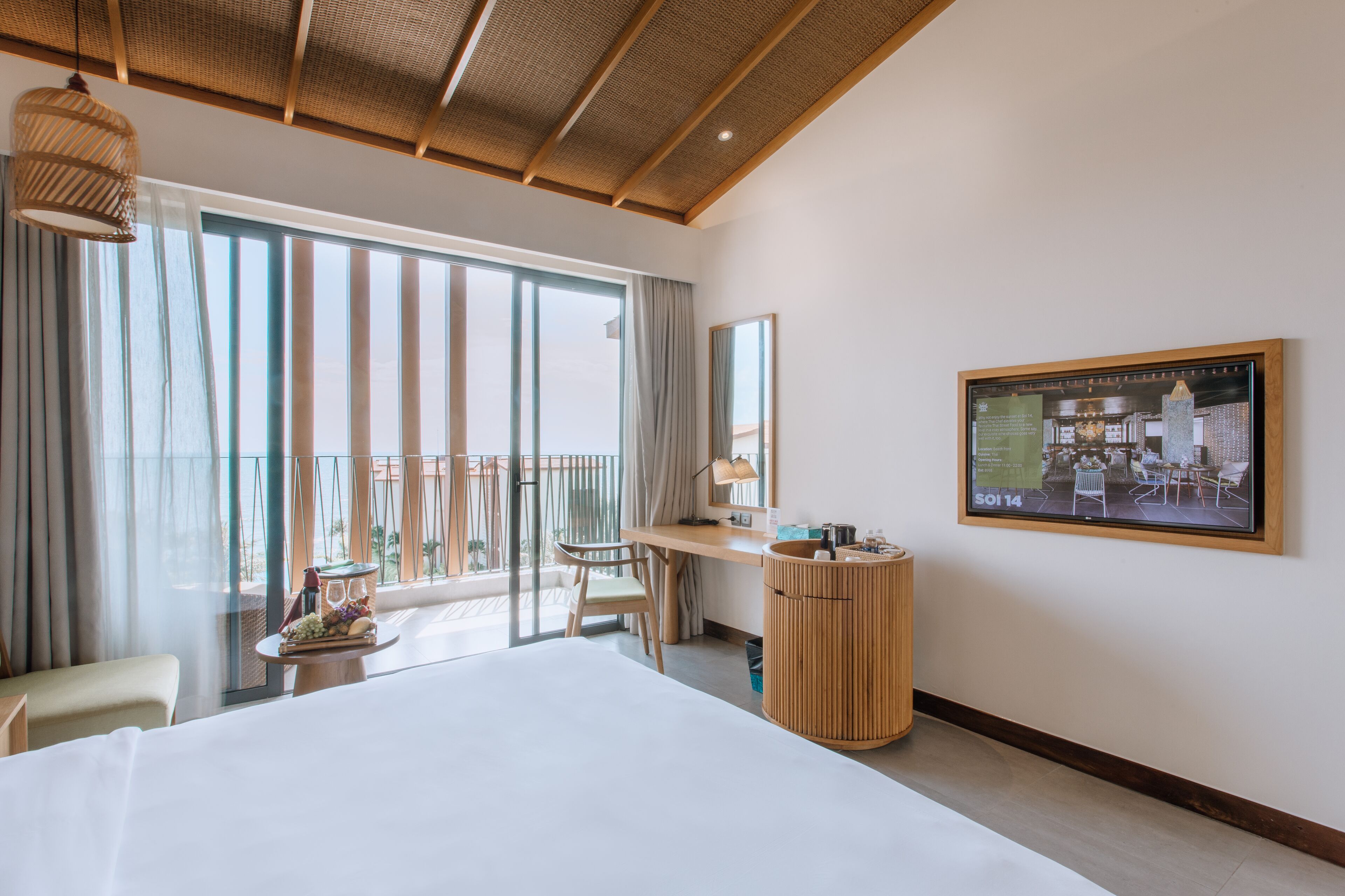 A hotel room with a large bed, a desk, a chair, a small table with a fruit basket, a waste bin, and a large window with curtains.