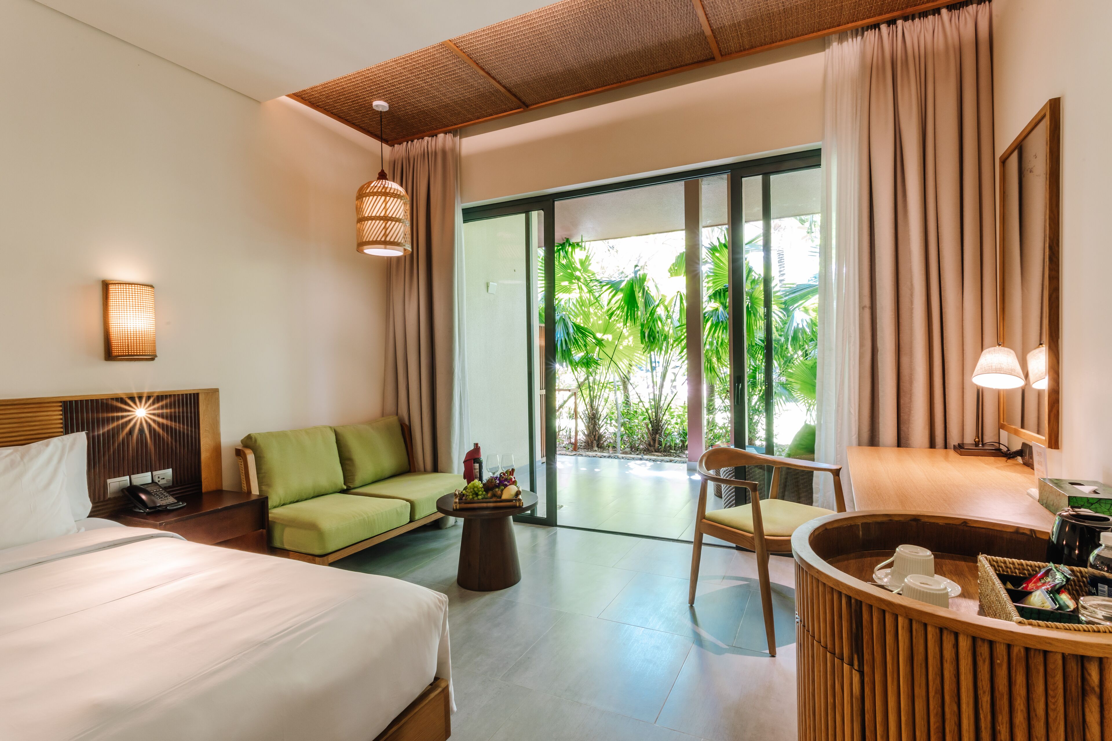A hotel room with a bed, a green sofa, a wooden coffee table with fruit, a wooden chair, and a view of greenery outside.