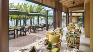 A restaurant with outdoor seating overlooking a beach, featuring wooden tables, chairs, and umbrellas. - Dusit Princess Moonrise Beach Resort (Phu Quoc)